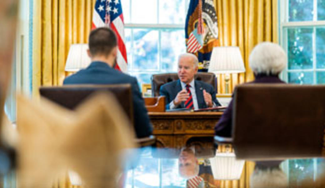 President Biden, sitting at his desk in the Oval Office, with his reflection in the foreground