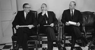 Walter W. Heller wearing glasses, Kermit Gordon has hand to chin, Luther Hodges looking at them both, all seated in blk chairs with papers in hand.