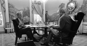 Sec. Clark Clifford (left) seated at table, speaking and gesturing as President Lyndon B. Johnson listens