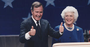 George H. W. Bush giving a thumbs up at a podium with Barbara Bush standing next to him 