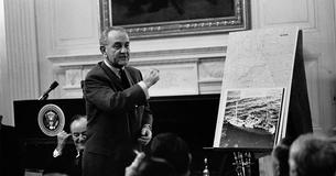 LBJ standing and talking in front of a photo of the USS Pueblo