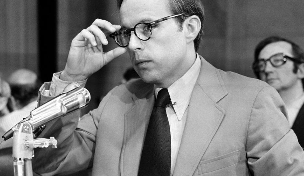 John Dean in front of a mic reaching up to touch his glasses