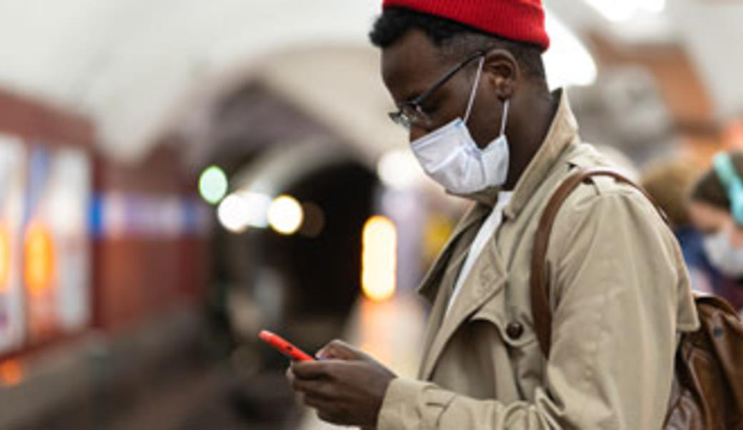 Man on subway platform with mask on looking at smartphone