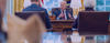 President Biden, sitting at his desk in the Oval Office, with his reflection in the foreground